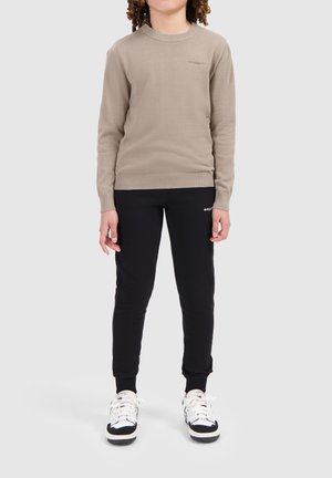 Beige gebreide trui met geribbelde ronde hals, gecombineerd met zwarte sweatpants met een logo. Model draagt witte en zwarte sneakers.