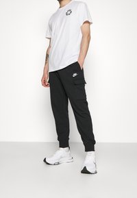 Vit bomullströja med svart grafik på bröstet, tillsammans med svarta joggerbyxor med sidoficka och vit Nike-logga. Vita sneakers.