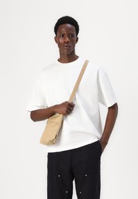 White oversized t-shirt, dark pants with pockets, holding a small beige crossbody bag. Simple design and neutral colors.