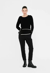 Črn pleten pulover z belim poudarkom na robu, združen s črnimi stožčastimi hlačami in črnimi škornji z vezalkami. Modelka pozira z rokami na bokih.