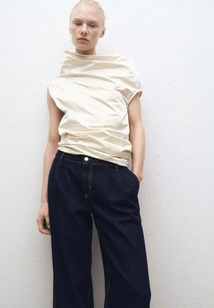 Cremfarbener Oberteil mit drapiertem Design, hohem Kragen, kombiniert mit dunkelblauen weiten Jeans mit fünf Taschen und Knopfverschluss.