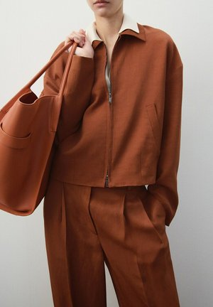 Persona con chaqueta color óxido y pantalones plisados a juego, sosteniendo un gran bolso tote del mismo color sobre el hombro.