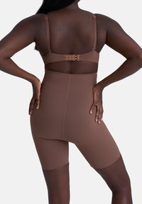 Vrouw in bruine hoge shaping shorts en bijpassende beha, staand met handen op de heupen tegen een effen lichte achtergrond.