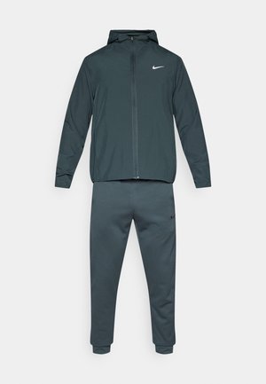 Σαλοπέτα Nike, που αποτελείται από μια ζακέτα με κουκούλα και εφαρμοστά παντελόνια. Κατασκευασμένη από σκούρο γκρι ύφασμα με λεία υφή και λευκά λογότυπα.