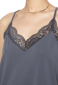 Camisole grise avec des accents en dentelle le long de l'encolure en V et des bretelles spaghetti, fabriquée à partir d'un tissu lisse avec un léger brillant.