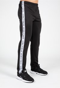Zwarte joggers met witte "GORILLA WEAR" branding langs de zijkant. Gemaakt van gladde stof, voorzien van zijzakken met rits en een elastische tailleband.