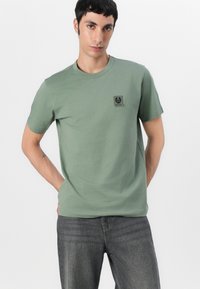 Tricou din bumbac verde cu gât rotund și mâneci scurte, având un patch cu logo negru pe piept. Asortat cu blugi gri închis.