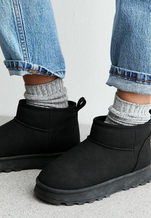 Stivali invernali neri indossati con calze in maglia grigia e jeans azzurri arrotolati che poggiano su un pavimento grigio.