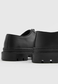 Chaussures en cuir noir avec une texture lisse, une forme arrondie et une semelle épaisse et robuste avec un motif de traction visible.