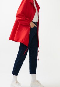 Rød, letvægts regnjakke med trykknapper, lange ærmer og snoretræk i kanten, parret med mørkeblå denimjeans og hvide sneakers.