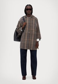 Sobretudo de lã à linha em castanho com realces em bege e laranja, apresentando uma silhueta ampla, fecho de botão e grandes bolsos. Combinado com jeans escuros.