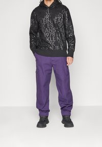 Black hoodie with a textured logo print, paired with loose-fitting, purple cargo pants featuring side pockets. Footwear is black.