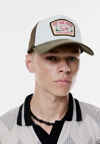 Baseballcap mit einem weißen Vorderpanel, braunem Netzrückenteil, runder Applikation mit "DRY OAK CREEK" und einem grünen Schild. Halskette aus schwarzen Perlen.