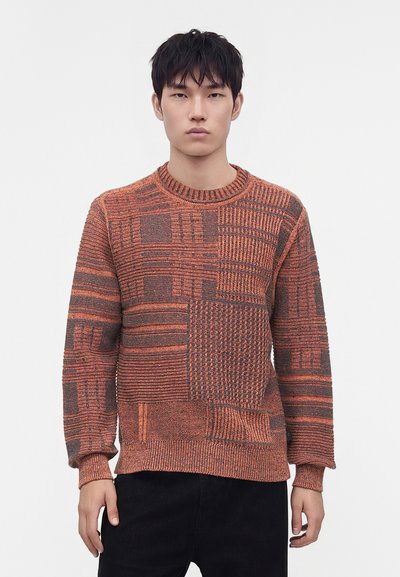 Jeune homme portant un pull en maille à motif patchwork orange et marron et un pantalon noir, debout devant un fond blanc uni.