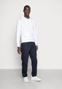 Witte hoodie met een voorzak, gecombineerd met marineblauwe broek en witte sneakers, op een lichtgrijze achtergrond.
