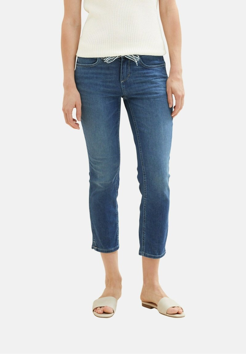 Mujer vestida con jeans de tiro corto en denim azul y cintura con cordón, blusa de punto en color blanco roto y sandalias planas beige, contra un fondo liso.