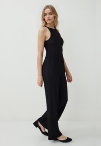 Vrouw die staat in een zwart mouwloos topje, textuur brede zwarte broek en zwarte flats tegen een eenvoudige lichte achtergrond.