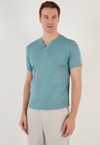 Tričko s krátkym rukávom a výstrihom do V v teal farbe, vyrobené z mäkkej zmesi bavlny. Obsahuje tri dekoratívne gombíky na krku a má voľný strih.