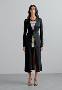 Larga chaqueta negra hecha de tela texturizada, con un cierre de botones en la parte delantera y mangas largas. Llevada sobre un vestido colorido con estampado.