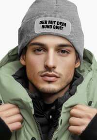 Gray knit beanie with a rectangular white patch and black text. The individual wears a green puffer jacket over a black layers.