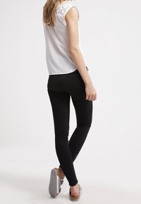 Blusa bianca senza maniche con dettagli in pizzo sulle spalle, abbinata a jeans skinny neri e sandali beige con suola testurizzata.