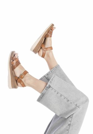 Sandalias de cuero marrón con correas perforadas y hebilla. Tienen una suela de plataforma gruesa con una textura similar a la madera, que se combina con unos pantalones vaqueros grises.