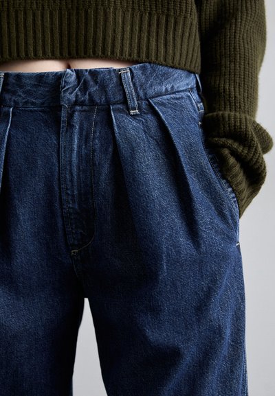 Højtragtede blå denimbukser med plisseringer, struktureret stof og en talje med bæltestropper. Sammen med en olivengrøn sweater.
