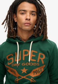 Sudadera verde con el texto "SUPER DRY GOODS" en naranja. Presenta cordones crema y un diseño texturizado. Camisa de cuadros visible debajo.