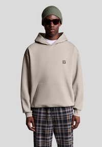 Felpa beige con tasca frontale e logo, indossata sopra una t-shirt bianca. Abbinata a pantaloni a quadri grigi e neri e a un berretto verde a coste.