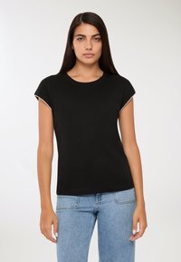 Młoda kobieta z długimi ciemnymi włosami, ubrana w czarny top z krótkim rękawem wykończonym perłowym brzegiem i jasnoniebieskie dżinsy, stojąca na tle białym.