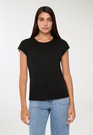 Giovane donna con lunghi capelli scuri, che indossa una maglietta nera a maniche corte con maniche bordate di perle e jeans azzurri, in piedi contro uno sfondo bianco.