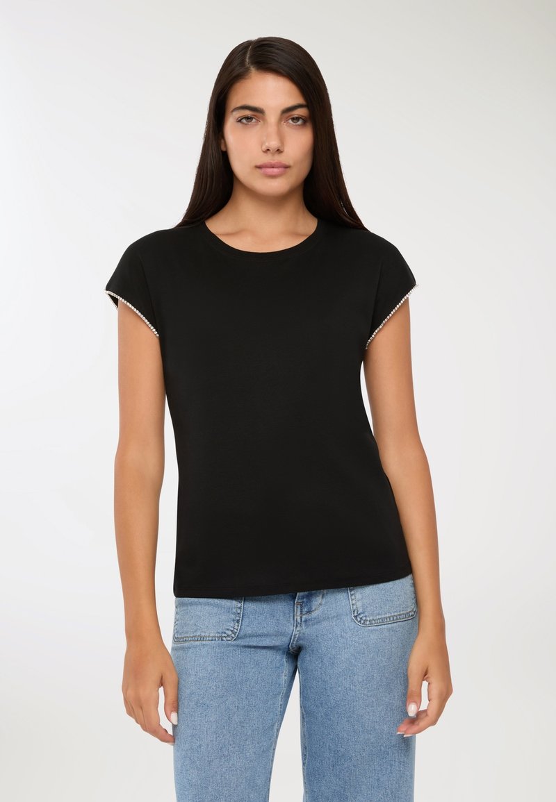 Młoda kobieta z długimi ciemnymi włosami, ubrana w czarny top z krótkim rękawem wykończonym perłowym brzegiem i jasnoniebieskie dżinsy, stojąca na tle białym.