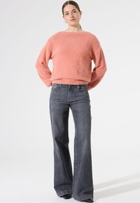 Rosa, samtiger Pullover mit weitem Rundhalsausschnitt, kombiniert mit hoch taillierten, dunkelgrauen Jeans. Der Pullover hat lange Ärmel und eine lockere Passform.