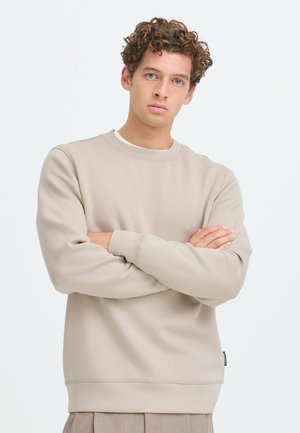 Jonge man met krullend haar, gekleed in een beige sweatshirt en beige broek, staand met gekruiste armen tegen een effen lichte achtergrond.