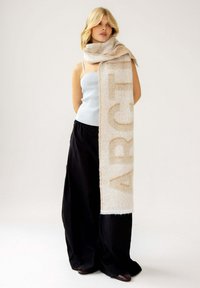 Beige sjaal met een zachte textuur, voorzien van een groot, lichtbruin logo. Model draagt een lichtblauw top en zwarte wijde broek.