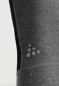 Grå atletiska leggings med ett texturerat tyg, med svarta accenter och ett cirkulärt logomönster i ljusgrått nära underbenet.