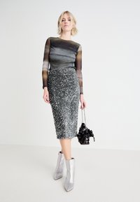 Crna sequin pencil suknja uparena s prozirnom, višebojnom bluzom dugih rukava. Model nosi srebrne metalik gležnjače i drži crnu torbicu.