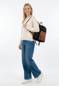 Smiling woman with long blonde hair wearing a cream sweater and jeans, carrying a black and brown backpack over one shoulder.