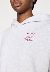 Siva kapucarica iz mehkega blaga z bordo rdečim logotipom, na katerem je napisano "ADIDAS three stripes STUDIO" v stilizirani pisavi.
