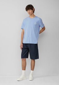 Lichtblauwe katoenen t-shirt met een kleine grafische afbeelding op de linkerborst, gecombineerd met donkere denimshorts en witte sneakers. Effen achtergrond.