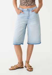 Culottes de mezclilla azul claro con un corte suelto, cintura a media altura, bolsillos delanteros y detalles de dobladillo sin rematar. Combinados con sandalias de tiras color marrón claro.