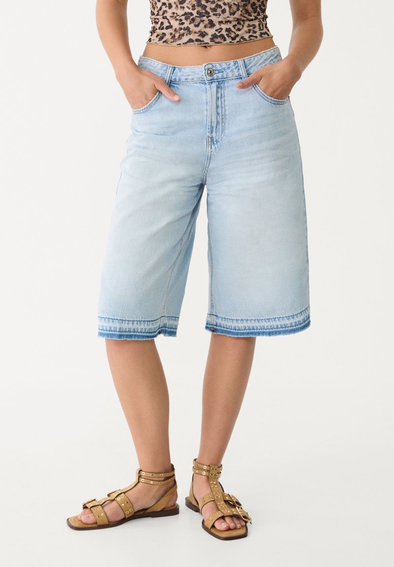 Culottes de mezclilla azul claro con un corte suelto, cintura a media altura, bolsillos delanteros y detalles de dobladillo sin rematar. Combinados con sandalias de tiras color marrón claro.