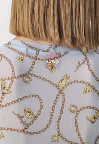 Valaistusverkko ylhäällä vaaleansinisessä, jossa on kultaketju ja kuvioita, mukaan lukien sydämiä, saappaita ja tähtiä. Logo "HUGO" kullassa selässä.