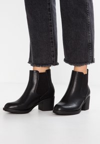 Botas de cuero negro hasta el tobillo con paneles laterales elásticos, punta redondeada y tacones en bloque. Suela de goma texturizada y jeans con dobladillo deshilachado son visibles.