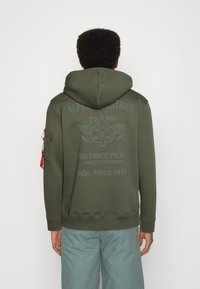 Felpa verde oliva con cappuccio, caratterizzata da motivi e testi di aviazione stampati sul retro. Dotata di una tasca laterale e polsini a coste.