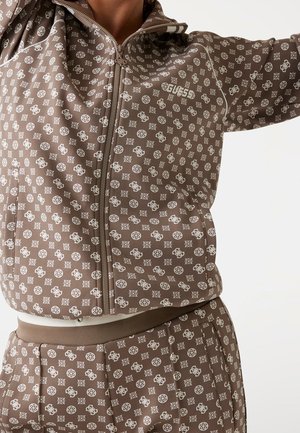 Veste marron zippée et pantalon avec motif du logo blanc Guess sur une personne avec le bras levé sur fond blanc.