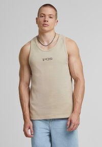 Tan geribbelde tanktop met een ronde hals en een logo in het midden. Draagt samen met lichtblauwe jeans, die een getailleerd ontwerp tonen.