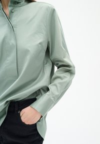Hellgrünes Langarmhemd mit Stehkragen und einer kleinen Knopfleiste vorne. Der Stoff wirkt glatt; kombiniert mit schwarzen Hosen.