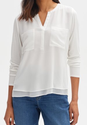 Vrouw draagt een wit blouse met lange mouwen en twee borstzakken aan de voorkant, gecombineerd met een blauwe spijkerbroek, tegen een effen lichte achtergrond.