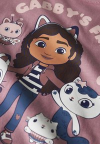 Chemise rose présentant une fille de dessin animé avec des cheveux bouclés foncés, portant une chemise rayée et un sweat à capuche marron, entourée de personnages d'animaux fantaisistes.